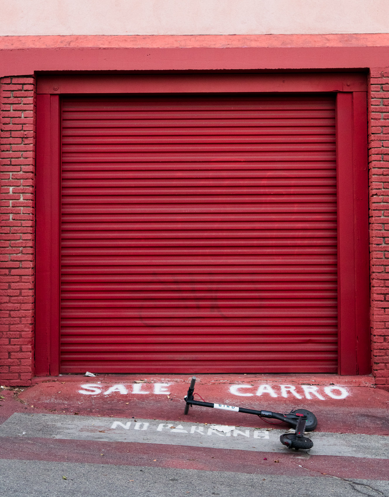 A Bird scooter lies in front of a red garage with a no parking sign in a Venice Beach alley.