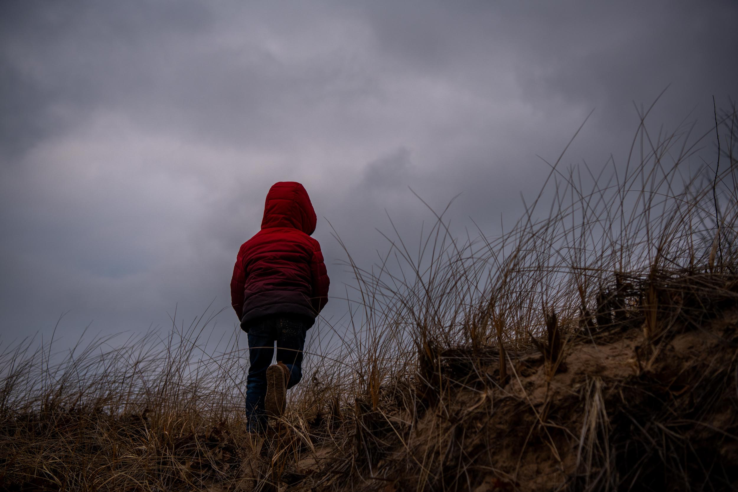 A boy hikes up a grassy dune wearing a red coat under cloudy skies in Michigan.