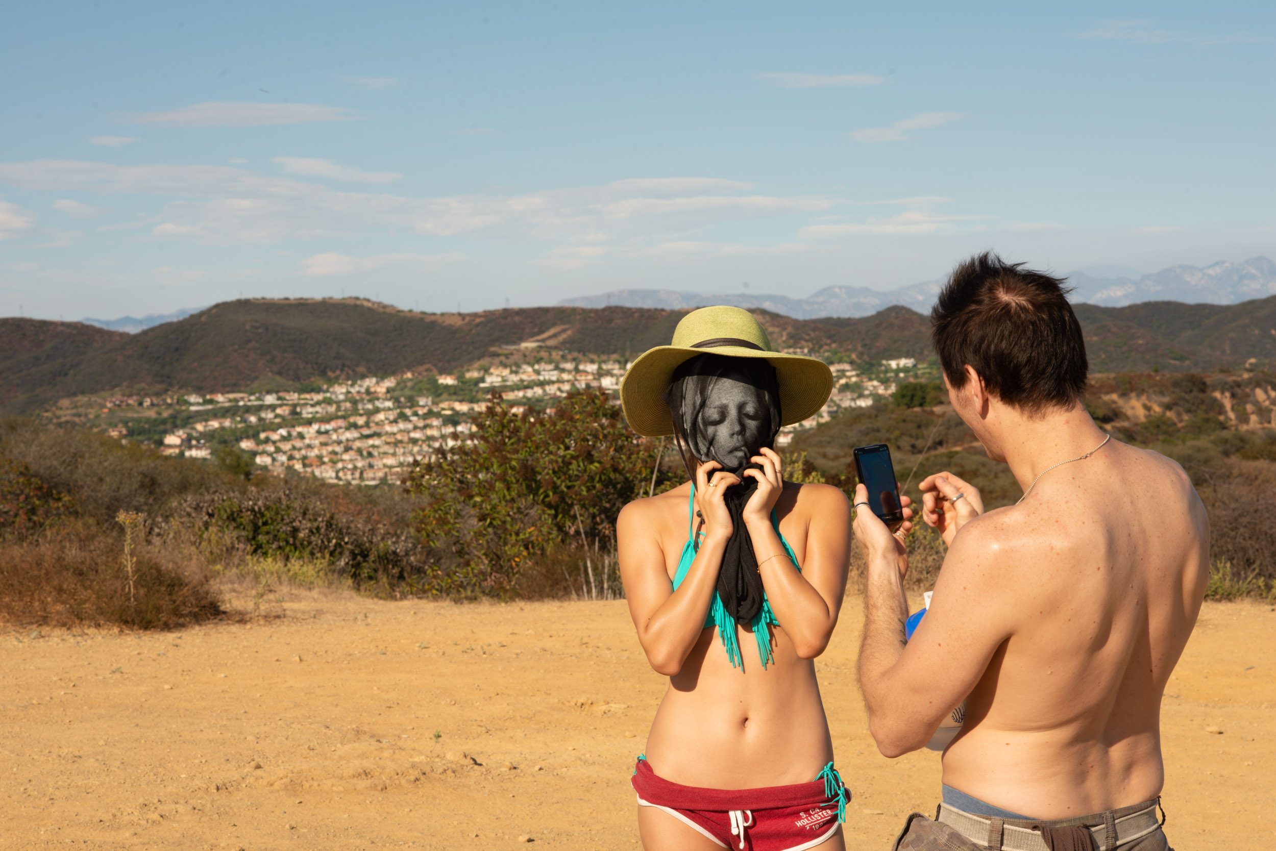 A man takes a picture of a woman holding a teeshirt mask over her face hiking in Malibu, Los Angeles.