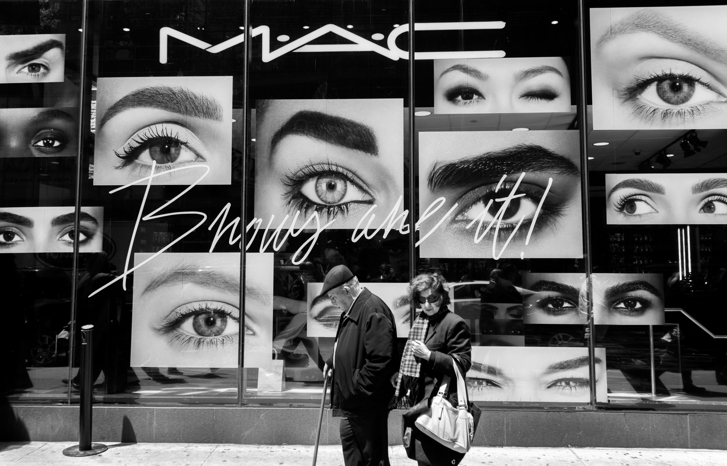 An older couple walks on a Manhattan sidewalk in front of pictures of large eyes at Mac Cosmetics.