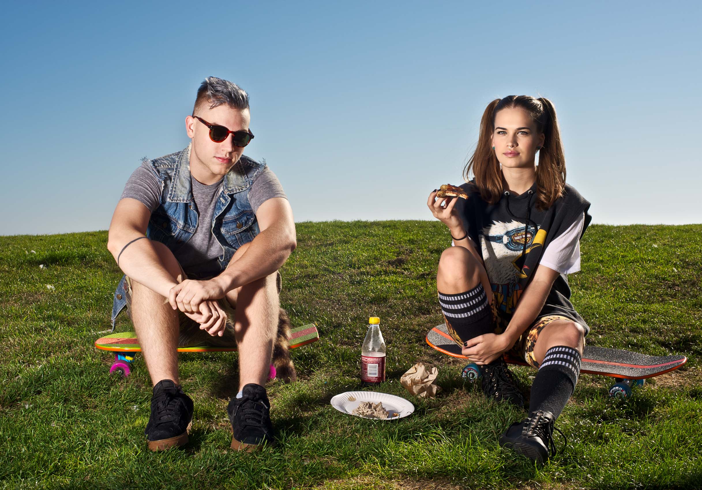 Portrait of a young man and woman eating pizza on grass along the Venice Beach boardwalk.